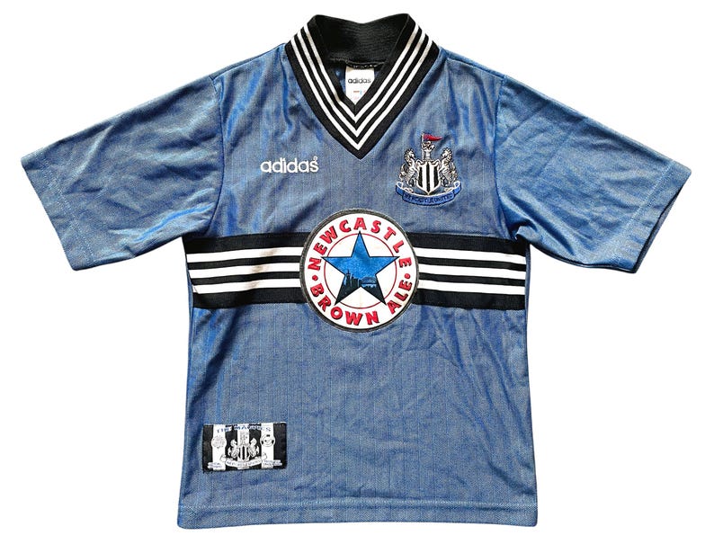 May include: A vintage blue soccer jersey with black and white accents. The jersey features the Adidas logo, a Newcastle United crest, and a "Newcastle Brown Ale" patch. The collar and sleeves have black and white stripes.