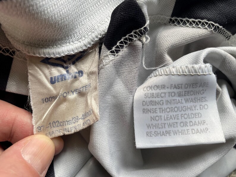 May include: White and black striped soccer jersey with a white tag that reads "100% POLYESTER 97-102cms/38-4" and a white tag that reads "COLOUR - FAST DYES ARE SUBJECT TO BLEEDING DURING INITIAL WASHES. RINSE THOROUGHLY. DO NOT LEAVE FOLDED WHILST WET OR DAMP RE-SHAPE WHILE DAMP"