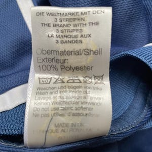 Op de afbeelding: Een wit label met blauwe tekst op een blauwe achtergrond. Het label luidt "Die Weltmarke mit den 3 Streifen", "The Brand with the 3 Stripes", "La Marque aux 3 Bandes", "Obermaterial/Shell Exterieur: 100% Polyester". Het label bevat ook wasinstructies in het Duits, Engels en Italiaans.