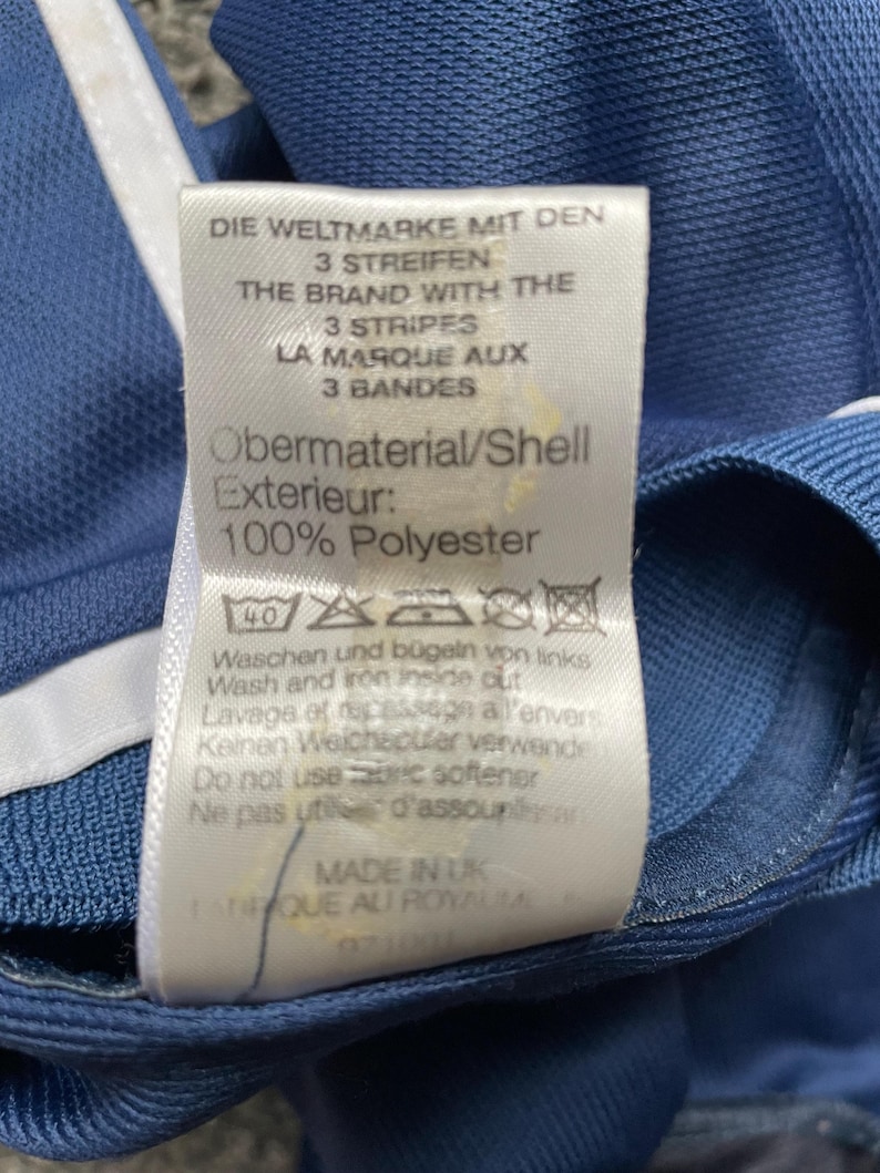Op de afbeelding: Een wit label met blauwe tekst op een blauwe achtergrond. Het label luidt "Die Weltmarke mit den 3 Streifen", "The Brand with the 3 Stripes", "La Marque aux 3 Bandes", "Obermaterial/Shell Exterieur: 100% Polyester". Het label bevat ook wasinstructies in het Duits, Engels en Italiaans.
