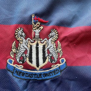 May include: Close-up of a blue, red, and black striped fabric. The fabric features an embroidered crest with two seahorses, a flag, and the words "NEWCASTLE UNITED" in a blue banner. The crest is white, black, and gold.