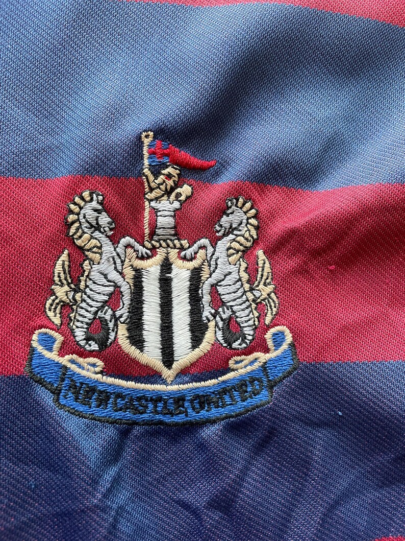 May include: Close-up of a blue, red, and black striped fabric. The fabric features an embroidered crest with two seahorses, a flag, and the words "NEWCASTLE UNITED" in a blue banner. The crest is white, black, and gold.
