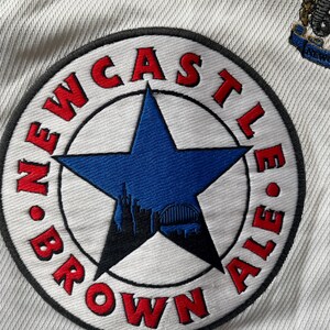 Pu&ograve; includere: Una toppa bianca con un cerchio nero e testo rosso che recita "NEWCASTLE BROWN ALE". All'interno del cerchio c'&egrave; una stella blu a cinque punte con una silhouette nera di uno skyline cittadino.