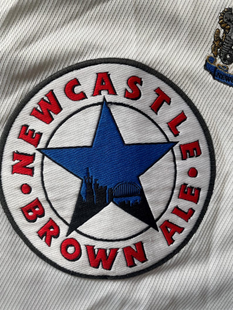 Pu&ograve; includere: Una toppa bianca con un cerchio nero e testo rosso che recita "NEWCASTLE BROWN ALE". All'interno del cerchio c'&egrave; una stella blu a cinque punte con una silhouette nera di uno skyline cittadino.