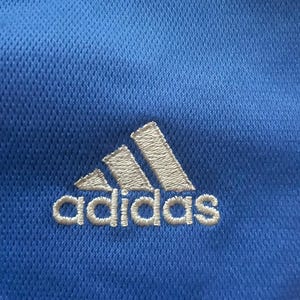 K&ouml;nnte beinhalten: Nahaufnahme eines blauen Sportshirts mit dem Adidas-Logo, wei&szlig; gestickt. Das Logo zeigt drei schr&auml;ge Streifen &uuml;ber dem Markennamen. Der Stoff hat ein strukturiertes, atmungsaktives Gewebe.
