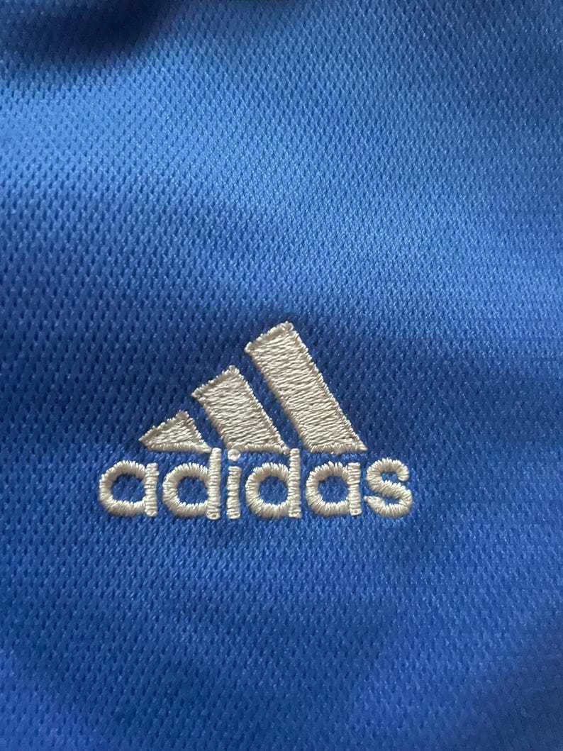 K&ouml;nnte beinhalten: Nahaufnahme eines blauen Sportshirts mit dem Adidas-Logo, wei&szlig; gestickt. Das Logo zeigt drei schr&auml;ge Streifen &uuml;ber dem Markennamen. Der Stoff hat ein strukturiertes, atmungsaktives Gewebe.