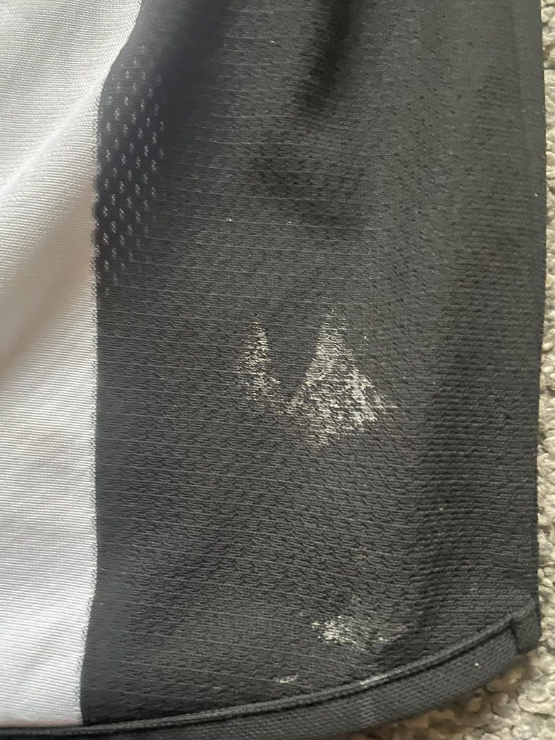 Puede incluir: Primer plano de una camiseta deportiva en blanco y negro. La camiseta tiene un panel blanco en el lado izquierdo y un panel negro en el lado derecho. Hay manchas blancas en el panel negro. La camiseta est&aacute; hecha de una tela transpirable y texturizada.