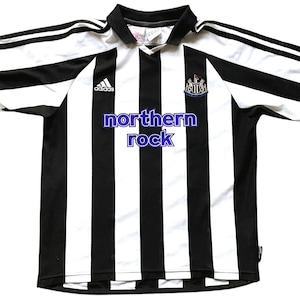 Pode incluir: Camisola de futebol de riscas pretas e brancas com o logotipo da Adidas e o bras&atilde;o do Newcastle United. O texto "northern rock" est&aacute; impresso na frente da camisola.