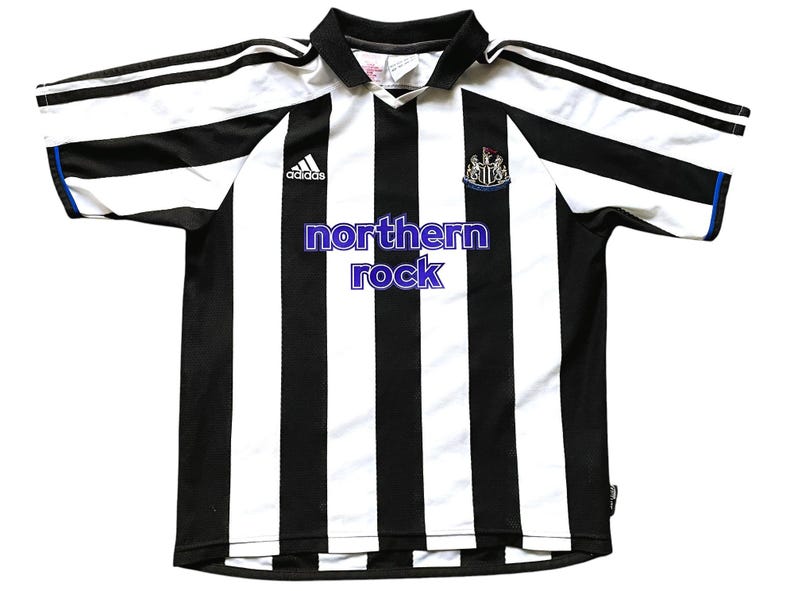 Pode incluir: Camisola de futebol de riscas pretas e brancas com o logotipo da Adidas e o bras&atilde;o do Newcastle United. O texto "northern rock" est&aacute; impresso na frente da camisola.