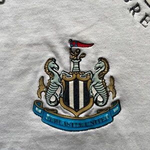 May include: White t-shirt with a black and white Newcastle United crest embroidered on the front. The crest features two seahorses, a shield with black and white stripes, and a banner that reads "Newcastle United".
