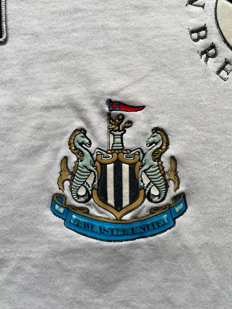 May include: White t-shirt with a black and white Newcastle United crest embroidered on the front. The crest features two seahorses, a shield with black and white stripes, and a banner that reads "Newcastle United".
