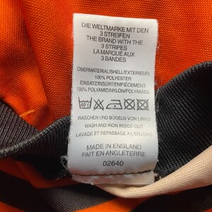 May include: A white tag with black text detailing the care instructions for a shirt. The tag reads "Die Weltmarke mit den 3 Streifen", "The Brand with the 3 Stripes", "La Marque aux 3 Bandes", "Obermaterial/Shell/Exterieur: 100% Polyester", "Einsatz/Insert/Empiecement: 100% Polyamid/Nylon/Polyamide", "Waschen und Bugeln von Links", "Wash and Iron Inside Out", "Lavage et Repassage a l'envers", "Made in England", "Fait en Angleterre", and "02640".