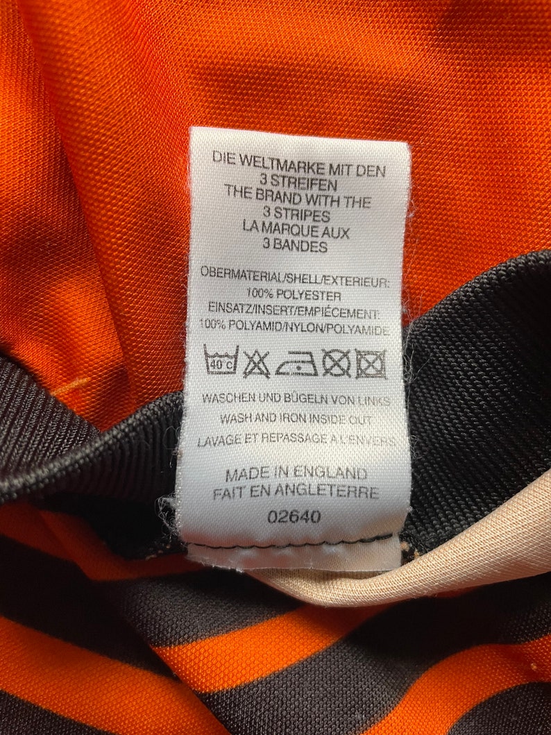 May include: A white tag with black text detailing the care instructions for a shirt. The tag reads "Die Weltmarke mit den 3 Streifen", "The Brand with the 3 Stripes", "La Marque aux 3 Bandes", "Obermaterial/Shell/Exterieur: 100% Polyester", "Einsatz/Insert/Empiecement: 100% Polyamid/Nylon/Polyamide", "Waschen und Bugeln von Links", "Wash and Iron Inside Out", "Lavage et Repassage a l'envers", "Made in England", "Fait en Angleterre", and "02640".