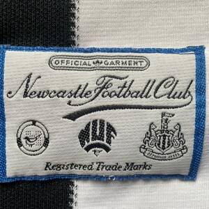 May include: A close-up of a label from a Newcastle Football Club jersey. The label is rectangular with a blue border and features the text "Newcastle Football Club" in cursive, along with several logos and the text "Registered Trade Marks."