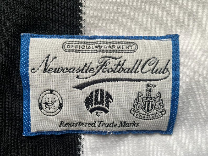 May include: A close-up of a label from a Newcastle Football Club jersey. The label is rectangular with a blue border and features the text "Newcastle Football Club" in cursive, along with several logos and the text "Registered Trade Marks."