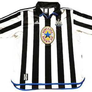 May include: A white and black striped soccer jersey with blue trim. The jersey has the Adidas logo on the left chest and a Newcastle Brown Ale logo on the right chest. The jersey has a short sleeve design.