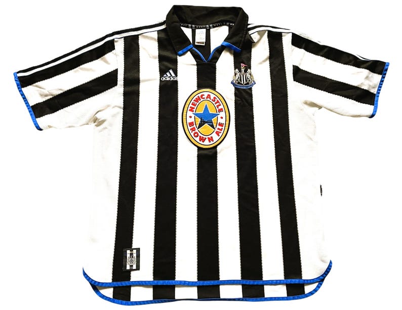 May include: A white and black striped soccer jersey with blue trim. The jersey has the Adidas logo on the left chest and a Newcastle Brown Ale logo on the right chest. The jersey has a short sleeve design.