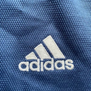 Op de afbeelding: Close-up van blauwe sportstof met het witte Adidas-logo geborduurd. Het Adidas-logo is een witte klaver met het woord "adidas" eronder in het wit geborduurd.