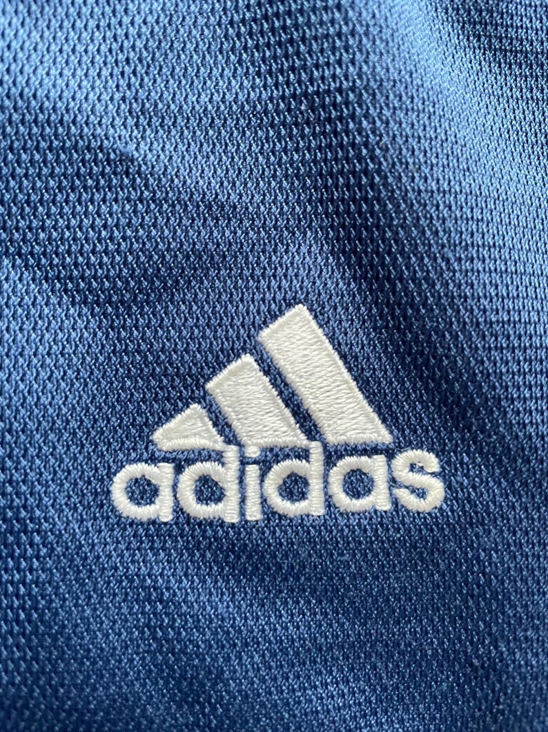 Op de afbeelding: Close-up van blauwe sportstof met het witte Adidas-logo geborduurd. Het Adidas-logo is een witte klaver met het woord "adidas" eronder in het wit geborduurd.