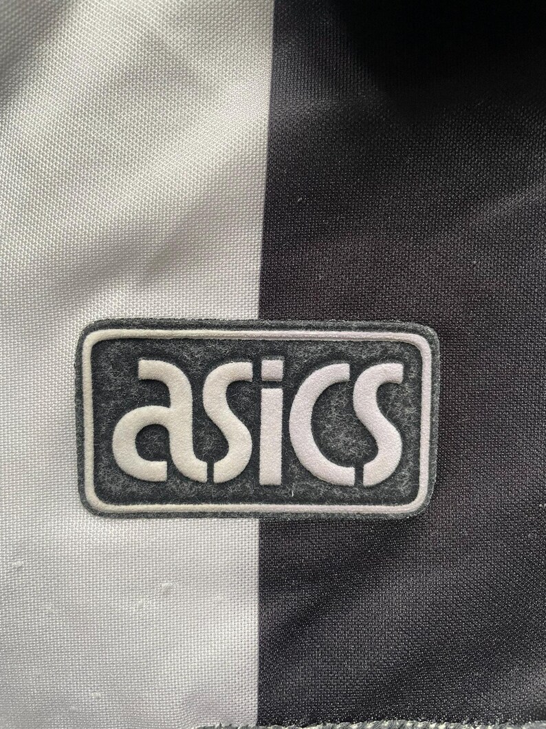 K&ouml;nnte beinhalten: Nahaufnahme eines schwarz-wei&szlig;en Sporttrikots mit dem Markennamen "asics" in wei&szlig;er Schrift auf einem grauen rechteckigen Aufn&auml;her. Das Trikot hat ein vertikales Streifendesign.
