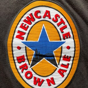 May include: A yellow, white, and red circular patch with the text "NEWCASTLE BROWN ALE" around the edge. A blue star with a cityscape silhouette inside is in the center.