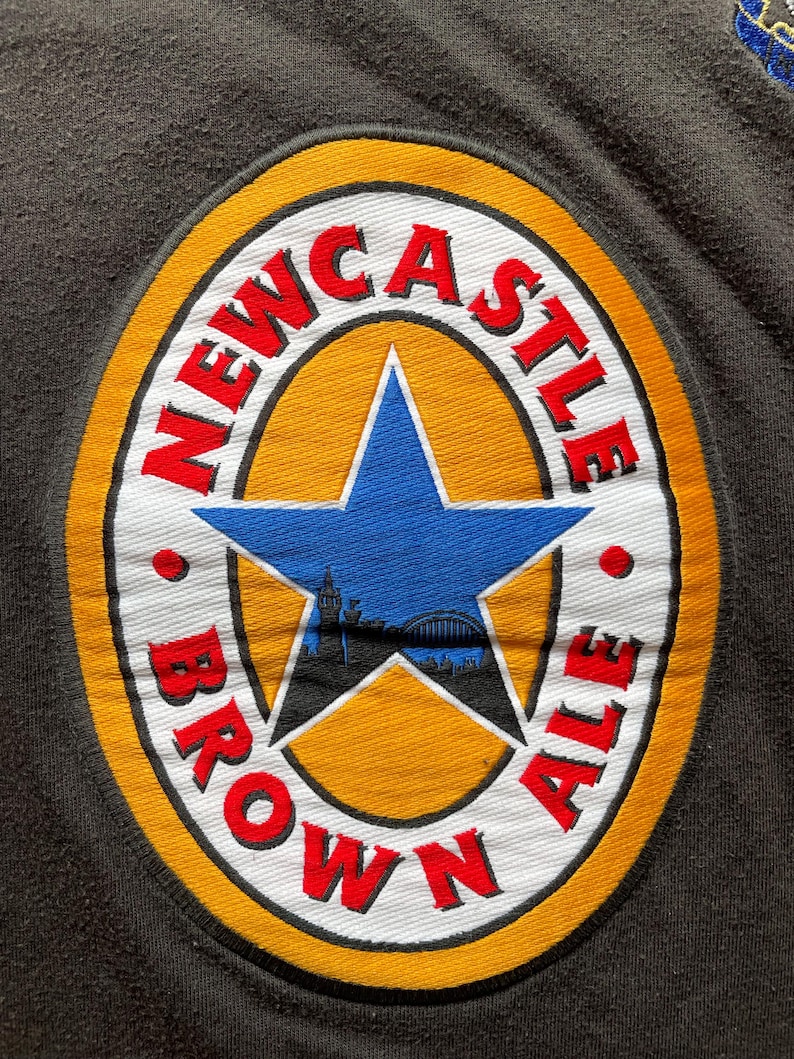 May include: A yellow, white, and red circular patch with the text "NEWCASTLE BROWN ALE" around the edge. A blue star with a cityscape silhouette inside is in the center.