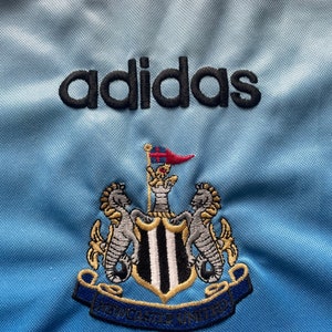 K&ouml;nnte beinhalten: Ein hellblaues Fu&szlig;balltrikot mit dem adidas-Logo und dem Wappen von Newcastle United. Das Wappen zeigt einen schwarz-wei&szlig;en Schild mit zwei Seepferdchen auf jeder Seite.