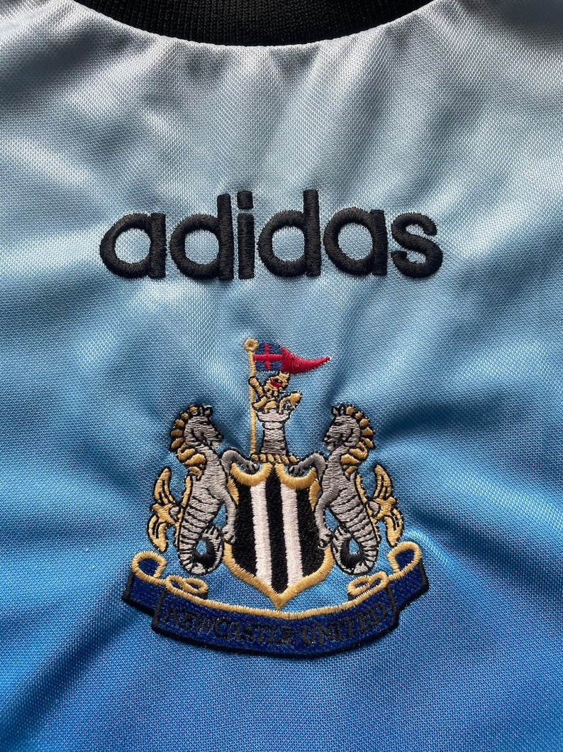 K&ouml;nnte beinhalten: Ein hellblaues Fu&szlig;balltrikot mit dem adidas-Logo und dem Wappen von Newcastle United. Das Wappen zeigt einen schwarz-wei&szlig;en Schild mit zwei Seepferdchen auf jeder Seite.