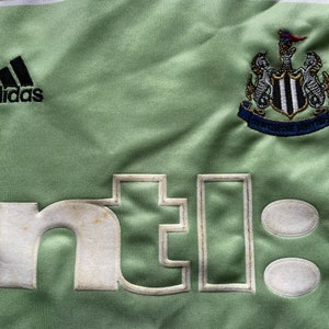 May include: A green soccer jersey with the Adidas logo, the Newcastle United crest, and the text "ntel" on the front.