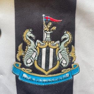 May include: Close-up of a black and white striped shirt with a Newcastle United crest. The crest features two seahorses, a shield with vertical stripes, and a banner with the team name. The shirt has a black collar.