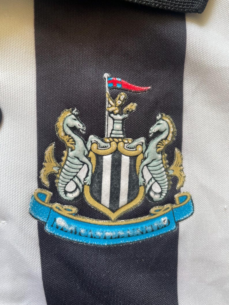 May include: Close-up of a black and white striped shirt with a Newcastle United crest. The crest features two seahorses, a shield with vertical stripes, and a banner with the team name. The shirt has a black collar.