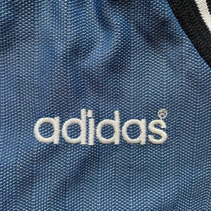 May include: Close-up of a blue fabric with the white embroidered Adidas logo. The fabric has a textured pattern. A black and white striped detail is visible in the upper right corner.