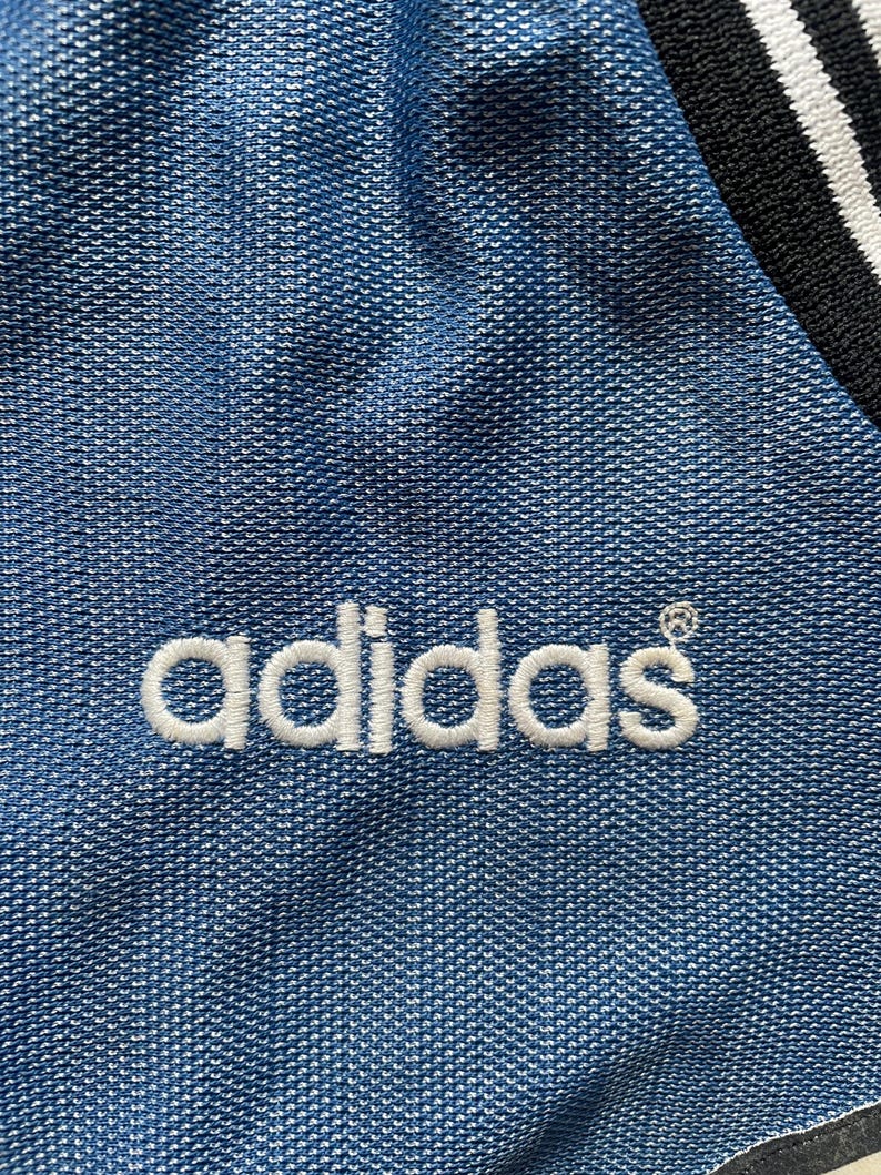 May include: Close-up of a blue fabric with the white embroidered Adidas logo. The fabric has a textured pattern. A black and white striped detail is visible in the upper right corner.