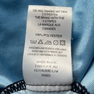 K&ouml;nnte beinhalten: Wei&szlig;es Pflegeetikett mit schwarzem Text und Symbolen. Der Text lautet "Die Weltmarke mit den 3 Streifen", "The Brand with the 3 Stripes", "La Marque aux 3 Bandes", "100% Polyester", "Waschen und B&uuml;geln von Links", "Wash and Iron Inside Out", "Lavage et Repassage a l'Envers", "Made in UK", "Fabrique au Royaume-Uni", "16593".