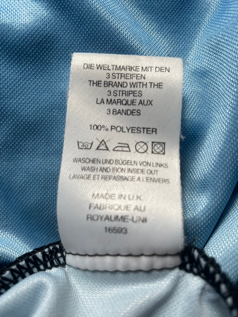K&ouml;nnte beinhalten: Wei&szlig;es Pflegeetikett mit schwarzem Text und Symbolen. Der Text lautet "Die Weltmarke mit den 3 Streifen", "The Brand with the 3 Stripes", "La Marque aux 3 Bandes", "100% Polyester", "Waschen und B&uuml;geln von Links", "Wash and Iron Inside Out", "Lavage et Repassage a l'Envers", "Made in UK", "Fabrique au Royaume-Uni", "16593".