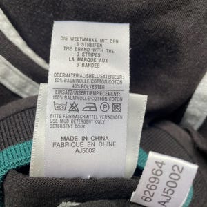May include: White tag with black text on a black shirt. The tag says "Die Weltmarke mit den 3 Streifen" and "The Brand with the 3 Stripes" and "La Marque aux 3 Bandes". The tag also says "Obermaterial/Shell/Exterieur: 60% Baumwolle/Cotton/Coton 40% Polyester" and "Einsatz/Insert/Empiecement: 100% Baumwolle/Cotton/Coton". The tag also has a care label with a 40 degree Celsius symbol, a hand wash symbol, a do not bleach symbol, a do not tumble dry symbol, and an iron symbol. The tag also says "Bitte Feinwaschmittel verwenden" and "Use Mild Detergent Only" and "Detergent Doux". The tag also says "Made in China" and "Fabrique en Chine" and "AJ5002".