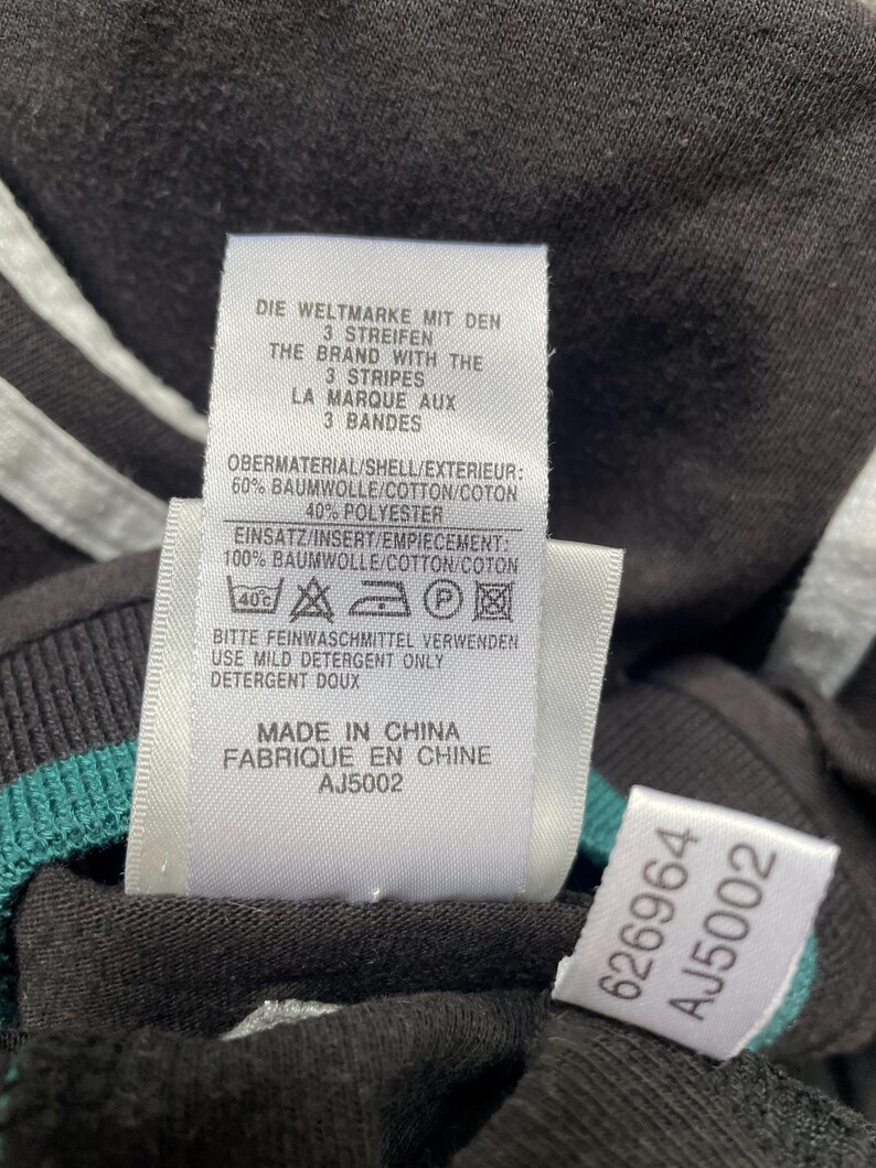 May include: White tag with black text on a black shirt. The tag says "Die Weltmarke mit den 3 Streifen" and "The Brand with the 3 Stripes" and "La Marque aux 3 Bandes". The tag also says "Obermaterial/Shell/Exterieur: 60% Baumwolle/Cotton/Coton 40% Polyester" and "Einsatz/Insert/Empiecement: 100% Baumwolle/Cotton/Coton". The tag also has a care label with a 40 degree Celsius symbol, a hand wash symbol, a do not bleach symbol, a do not tumble dry symbol, and an iron symbol. The tag also says "Bitte Feinwaschmittel verwenden" and "Use Mild Detergent Only" and "Detergent Doux". The tag also says "Made in China" and "Fabrique en Chine" and "AJ5002".