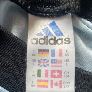 K&ouml;nnte beinhalten: Wei&szlig;es Etikett mit schwarzem Text und einem blauen Adidas-Logo. Das Etikett zeigt Gr&ouml;&szlig;enumrechnungen f&uuml;r verschiedene L&auml;nder, darunter Deutschland, Gro&szlig;britannien, die Vereinigten Staaten, die Schweiz, Kanada und Spanien. Das Etikett lautet "adidas" und enth&auml;lt die Gr&ouml;&szlig;en: D 164, GB Y, F XS, US L, I 14, CH 164, E 164, CDN LIG.