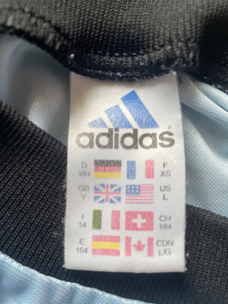 K&ouml;nnte beinhalten: Wei&szlig;es Etikett mit schwarzem Text und einem blauen Adidas-Logo. Das Etikett zeigt Gr&ouml;&szlig;enumrechnungen f&uuml;r verschiedene L&auml;nder, darunter Deutschland, Gro&szlig;britannien, die Vereinigten Staaten, die Schweiz, Kanada und Spanien. Das Etikett lautet "adidas" und enth&auml;lt die Gr&ouml;&szlig;en: D 164, GB Y, F XS, US L, I 14, CH 164, E 164, CDN LIG.
