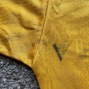 May include: Close-up of a mustard yellow fabric with a textured pattern. The fabric has dark stains near the seam. The sleeve of the garment is visible, with the fabric's texture and color prominent.