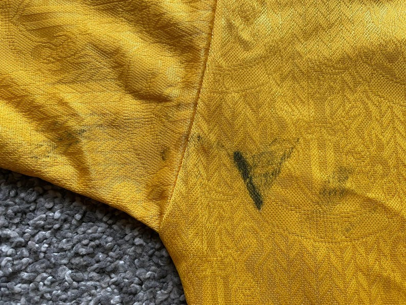 May include: Close-up of a mustard yellow fabric with a textured pattern. The fabric has dark stains near the seam. The sleeve of the garment is visible, with the fabric's texture and color prominent.