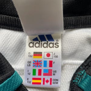 May include: White Adidas clothing tag with size chart in various languages and measurements. The tag is white with black text and a blue Adidas logo. The size chart includes sizes for Germany, Japan, Great Britain, Italy, the United States, Spain, and Canada.
