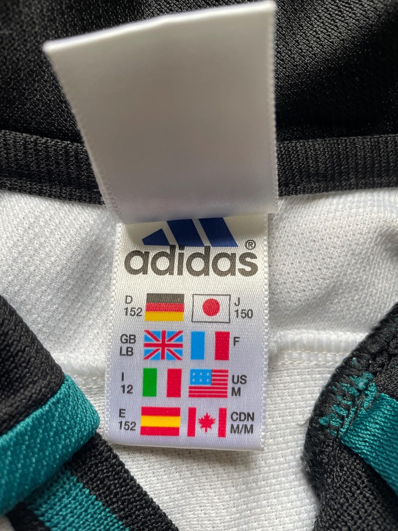 May include: White Adidas clothing tag with size chart in various languages and measurements. The tag is white with black text and a blue Adidas logo. The size chart includes sizes for Germany, Japan, Great Britain, Italy, the United States, Spain, and Canada.