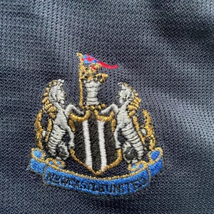 Puede incluir: Un escudo bordado en blanco y negro con un contorno dorado. El escudo presenta un escudo con rayas verticales blancas y negras, dos animales blancos a cada lado del escudo y una bandera roja y azul en la parte superior. El texto "NEWCASTLE UNITED" est&aacute; escrito en una cinta azul debajo del escudo.