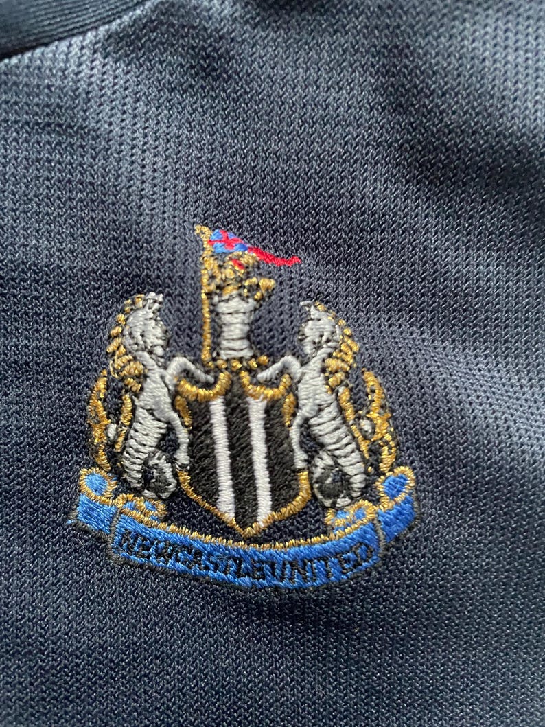 Puede incluir: Un escudo bordado en blanco y negro con un contorno dorado. El escudo presenta un escudo con rayas verticales blancas y negras, dos animales blancos a cada lado del escudo y una bandera roja y azul en la parte superior. El texto "NEWCASTLE UNITED" est&aacute; escrito en una cinta azul debajo del escudo.