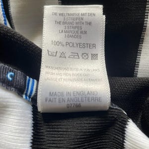 May include: Close-up of a tag with text in multiple languages, including English and French. The tag is white with black text and is attached to a black and white striped fabric. The tag reads "100% POLYESTER".