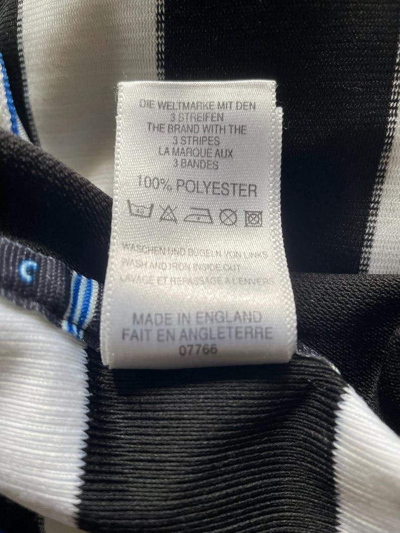 May include: Close-up of a tag with text in multiple languages, including English and French. The tag is white with black text and is attached to a black and white striped fabric. The tag reads "100% POLYESTER".