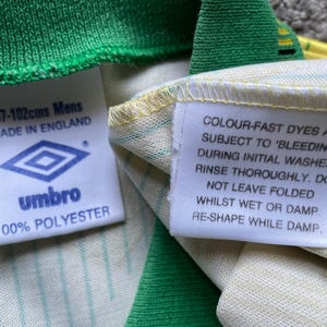 May include: White and green striped shirt with a white tag that says "COLOUR-FAST DYES ARE SUBJECT TO 'BLEEDING' DURING INITIAL WASHES. RINSE THOROUGHLY. DO NOT LEAVE FOLDED WHILST WET OR DAMP. RE-SHAPE WHILE DAMP." and a white tag with a logo and the text "97-102cms Mens MADE IN ENGLAND umbro 100% POLYESTER"