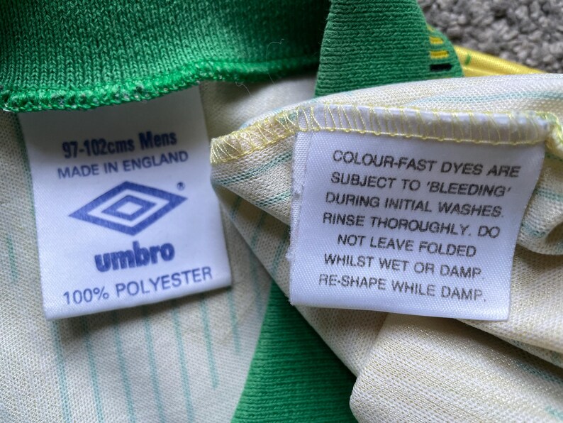 May include: White and green striped shirt with a white tag that says "COLOUR-FAST DYES ARE SUBJECT TO 'BLEEDING' DURING INITIAL WASHES. RINSE THOROUGHLY. DO NOT LEAVE FOLDED WHILST WET OR DAMP. RE-SHAPE WHILE DAMP." and a white tag with a logo and the text "97-102cms Mens MADE IN ENGLAND umbro 100% POLYESTER"