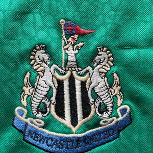 K&ouml;nnte beinhalten: Gesticktes Newcastle United Wappen auf gr&uuml;nem Stoff. Das Wappen zeigt zwei Seepferde, die einen Schild mit schwarz-wei&szlig;en Streifen flankieren. Ein Banner darunter lautet "NEWCASTLE UNITED". Oben befindet sich eine kleine Flagge.